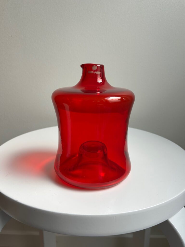 ruby-red glass bottle with a rounded, sculptural shape, photographed on a white stool