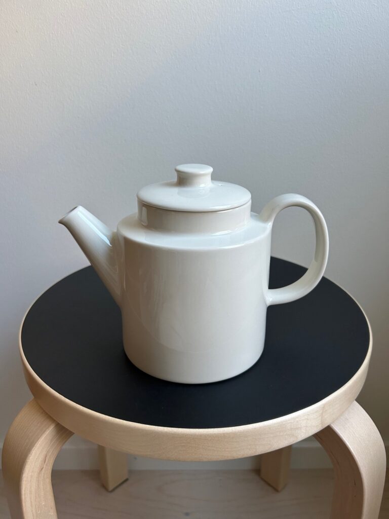 white ceramic teapot with a round body and straight spout, placed on a black-topped wooden stool