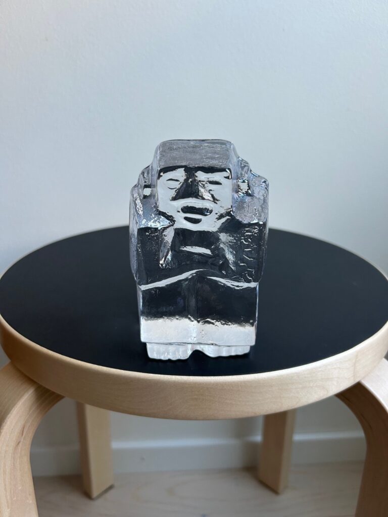 clear glass sculpture depicting a stylized human face, placed on a black round stool