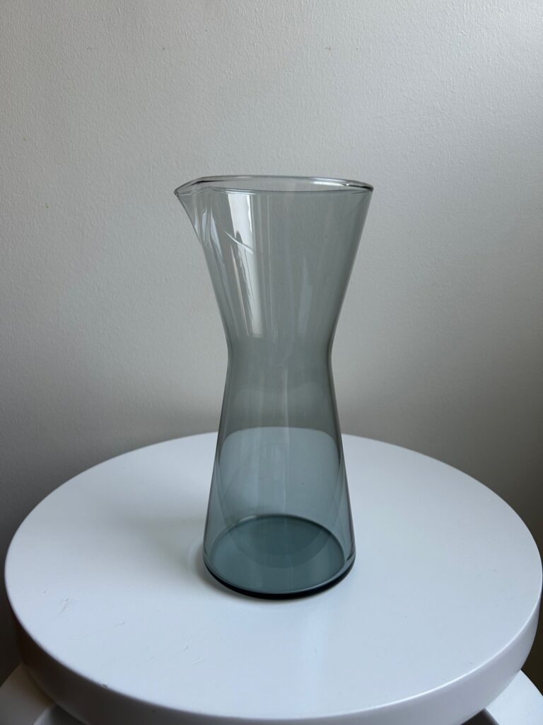 A tall grey glass carafe with a narrow neck, displayed on a white stool.