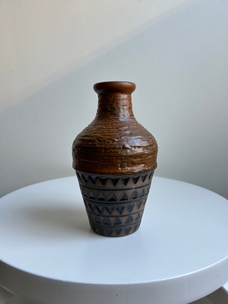 A small brown ceramic vase with geometric patterns, placed on a white stool.