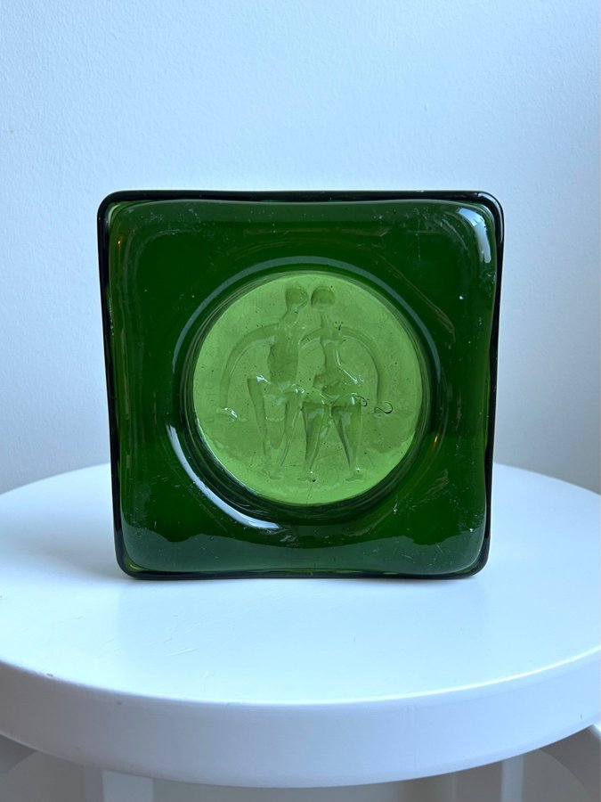 green square glass block with embossed figure motif, displayed on a white stool
